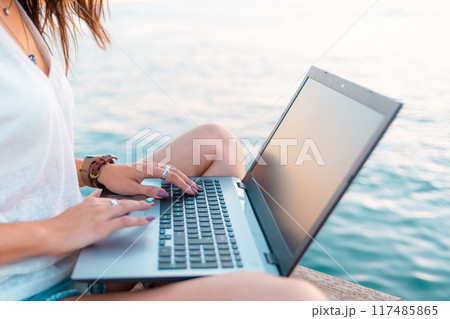 A woman works remotely on a laptop. Hands close-up. Sea in the background. The concept of freelancing A woman works remotely on a laptop. Hands close-up. Sea in the background. The concept of freelancing 117485865