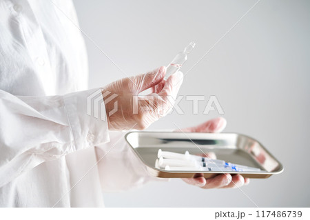 The doctor's gloved hands hold a tray with syringes and an ampoule with medicine. 117486739