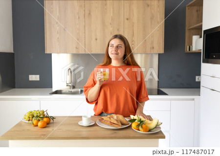 A happy 30s woman standing in a kitchen, holding a glass of fresh orange juice at home. Domestic life and leisure moments. Healthy diet. Young woman with glass of drink at table in kitchen. A happy 30s woman standing in a kitchen, holding a glass of fresh orange juice at home. Domestic life and leisure moments. Healthy diet. Young woman with glass of drink at table in kitchen. 117487017