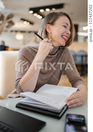 Young laughing woman working on laptop in cozy cafe with other people engaging in conversation in background. Concentrated study in co-working space. Education and resource searching concept Young laughing woman working on laptop in cozy cafe with other people engaging in conversation in background. Concentrated study in co-working space. Education and resource searching concept 117487262