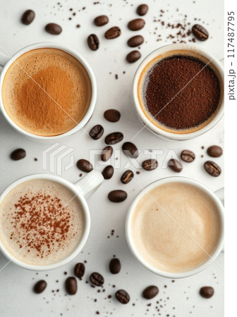 Three cups of coffee with ground coffee beans on a white surface and green leaves in the background 117487795