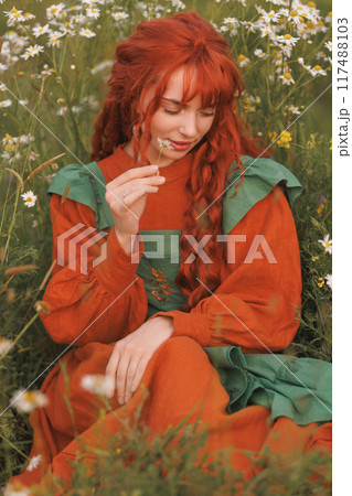 A shepherdess woman with red hair and a green dress sits in a field of flowers. A shepherdess woman with red hair and a green dress sits in a field of flowers. 117488103