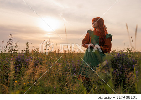 A shepherdess woman with red hair and a green dress sits in a field of flowers. 117488105
