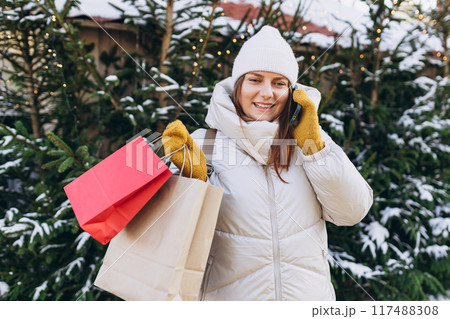 Young happy female in winter clothes with shopping bags standing in Christmas market with illuminated tree behind and speaking on mobile 117488308