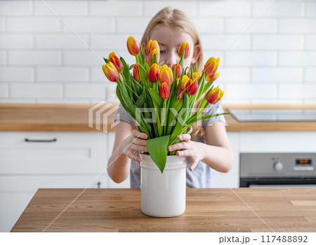 The girl puts a beautiful bouquet of red and yellow tulips in a white vase on a wooden countertop 117488892