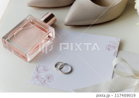 Bride's wedding essentials: heels, bouquet, rings, perfume, and invitation on a white table 117489019