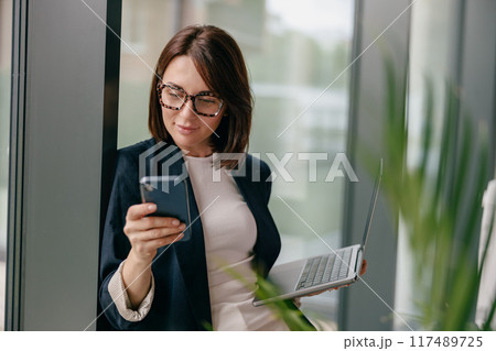 A young businesswoman in an office multitasking with a smartphone and laptop for work 117489725