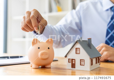 Close up hand of a businessman putting coin in a pink piggy bank, with a small house model on the table, saving or houise mortgage concept 117491675