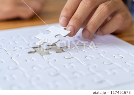 Close up hand of a man holding a white jigsaw puzzle piece trying to put it in an empty slot on the puzzle Close up hand of a man holding a white jigsaw puzzle piece trying to put it in an empty slot on the puzzle 117491678