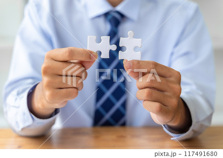 Close up hands of a businessman holding 2 jigsaw puzzle piecess trying to put them together Close up hands of a businessman holding 2 jigsaw puzzle piecess trying to put them together 117491680