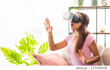 A girl in a pink shirt sits on a couch and experiences virtual reality with a VR headset on. She reaches out her hand in front of her as if she is interacting with something in the virtual world. A girl in a pink shirt sits on a couch and experiences virtual reality with a VR headset on. She reaches out her hand in front of her as if she is interacting with something in the virtual world. 117494426