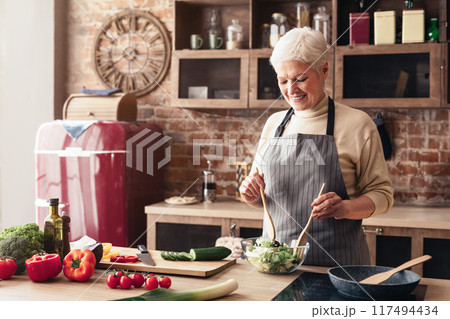 A senior woman in an apron smiles as she uses wooden spoons to mix a salad in a glass bowl on a kitchen counter. A red refrigerator is visible in the background, and other ingredients 117494434