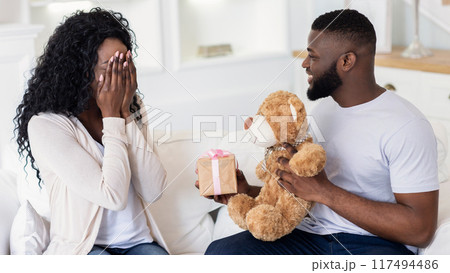 A man surprises a woman with a gift and a teddy bear while she covers her face with her hands in surprise. 117494486