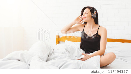 A woman is seated on a bed, wearing headphones and listening to music. She appears relaxed and focused on the music playing through the headphones. A woman is seated on a bed, wearing headphones and listening to music. She appears relaxed and focused on the music playing through the headphones. 117494537