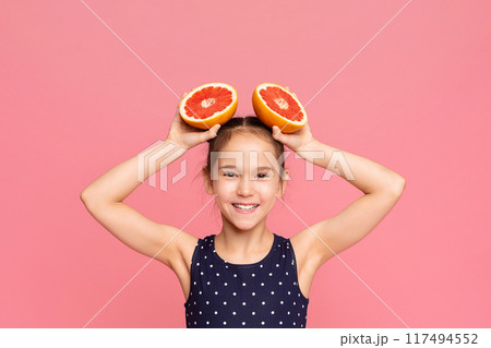A young girl is balancing two grapefruits on her head. She carefully holds them in place, displaying her skill and focus. The bright colors of the fruit contrast with her outfit A young girl is balancing two grapefruits on her head. She carefully holds them in place, displaying her skill and focus. The bright colors of the fruit contrast with her outfit 117494552