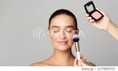 A woman is seen in close-up applying makeup to her face using a brush. She carefully contours her cheeks, brushes color onto her eyelids, and defines her eyebrows. 117494553