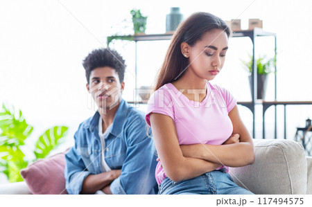 Black teen couple sits on a couch in a living room. They are both looking away from each other and appear to be upset. The girl has her arms crossed and is looking down Black teen couple sits on a couch in a living room. They are both looking away from each other and appear to be upset. The girl has her arms crossed and is looking down 117494575