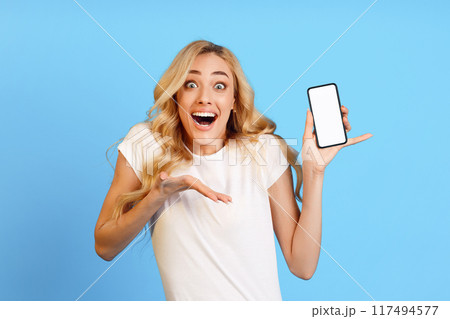 Amazed young woman with long, blonde hair wearing a white t-shirt standing in front of a blue background. She is holding a smartphone with a blank screen in her right hand 117494577