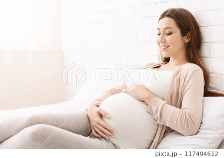 A pregnant woman is seated on a bed, her hand resting on her belly. She appears relaxed and comfortable as she prepares for rest or relaxation, copy space A pregnant woman is seated on a bed, her hand resting on her belly. She appears relaxed and comfortable as she prepares for rest or relaxation, copy space 117494612