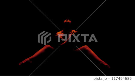 A woman is posed in a seated position, illuminated by red light. She has her arms crossed over her chest and is looking up towards the light source. The background is completely black 117494689