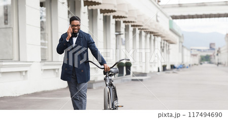 An African American businessman is riding a bicycle while talking on his phone. The scene is lively and contemporary, reflecting a busy yet balanced lifestyle. 117494690