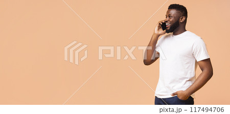 Young Black man talking on his phone while smiling and looking to the left. The man is wearing a white t-shirt and blue jeans and is standing against a beige background, copy space 117494706