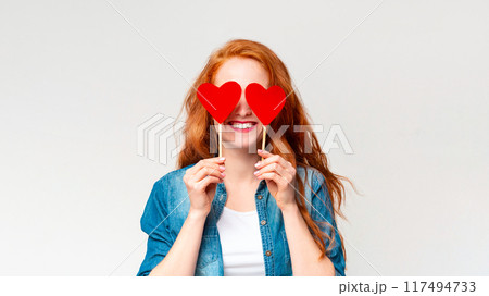 A cheerful woman with red hair and wearing a denim jacket stands against a plain white backdrop, holding two red heart-shaped cutouts in front of her eyes as if they were glasses A cheerful woman with red hair and wearing a denim jacket stands against a plain white backdrop, holding two red heart-shaped cutouts in front of her eyes as if they were glasses 117494733