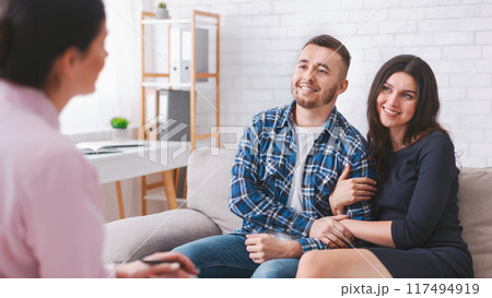 A young couple sits on a couch in a therapists office, listening intently to their counselor, who is out of focus in the foreground. 117494919