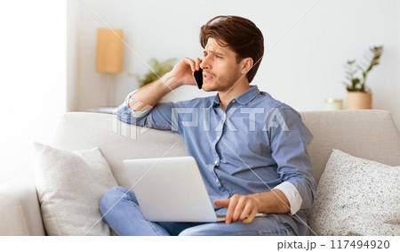 A man sits on a couch, talking on a phone while working on a laptop. 117494920