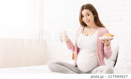 A pregnant woman is sitting comfortably in bed, smiling while holding a glass of milk and a pastry. She appears content and relaxed, showcasing a moment of calm and nourishment during her pregnancy. A pregnant woman is sitting comfortably in bed, smiling while holding a glass of milk and a pastry. She appears content and relaxed, showcasing a moment of calm and nourishment during her pregnancy. 117494927