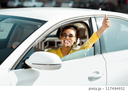 This image shows a happy black woman sitting in a white car, looking at the camera and giving a thumbs up out the window. Her bright yellow shirt and sunglasses add to her cheerful demeanor. 117494932
