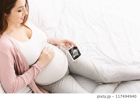 A pregnant woman is seen relaxing on a bed, holding an ultrasound image of her unborn baby. She is comfortably propped against a pillow and gently touching her belly, expressing joy and anticipation. A pregnant woman is seen relaxing on a bed, holding an ultrasound image of her unborn baby. She is comfortably propped against a pillow and gently touching her belly, expressing joy and anticipation. 117494940