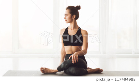 A woman is seated on a yoga mat, engaging in a yoga pose. She appears focused and balanced, showcasing flexibility and strength. The setting is simple and minimalistic 117494988