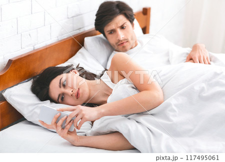 A woman is lying in bed, looking at her phone while a man, who is also in bed, watches her suspiciously. This image can be used to illustrate infidelity, mistrust, or relationship problems. A woman is lying in bed, looking at her phone while a man, who is also in bed, watches her suspiciously. This image can be used to illustrate infidelity, mistrust, or relationship problems. 117495061