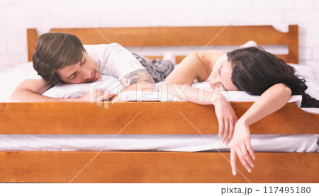 A man and a woman are lying in bed together, sleeping. The man is on his side, facing the woman. The woman is on her stomach, with her head resting on her arm. Both of them are asleep A man and a woman are lying in bed together, sleeping. The man is on his side, facing the woman. The woman is on her stomach, with her head resting on her arm. Both of them are asleep 117495180