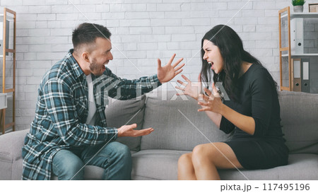 A couple is having a heated argument on a couch in their living room. The woman is sitting on the couch with her legs crossed and is gesturing wildly with her hands while yelling at the man. 117495196