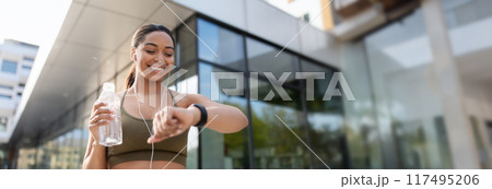 Young black woman looking at smartwatch, tracking fitness activity, listening to music in earphones, drinking water on training outdoors. Fit African American lady checking heart rate, copy space 117495206