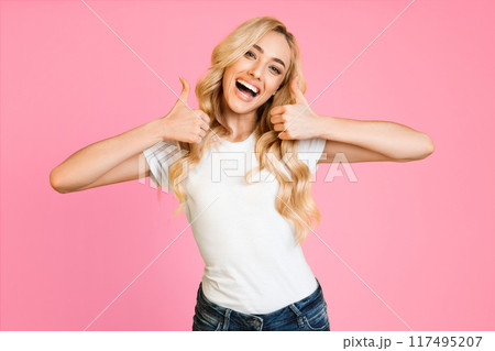 This image shows a happy blonde woman wearing a white t-shirt and blue jeans. She is giving a thumbs up with both hands and smiling. The background is a vibrant pink. 117495207
