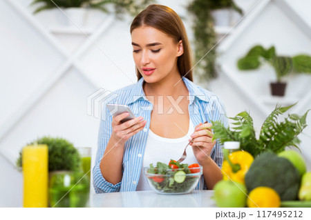A woman checks her phone while eating a fresh salad with a fork. The setting is a bright kitchen with plants in the background. 117495212