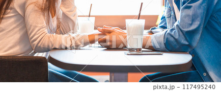 Two individuals, seated at a wooden table, are holding hands in a display of closeness and connection. The gesture suggests intimacy and mutual affection between the pair, cropped 117495240