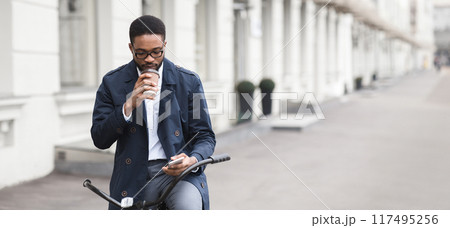 Black man professional is riding a bicycle to the office, carrying coffee and checking phone. The scene is modern and busy, capturing the essence of multitasking and an active start to the day. Black man professional is riding a bicycle to the office, carrying coffee and checking phone. The scene is modern and busy, capturing the essence of multitasking and an active start to the day. 117495256
