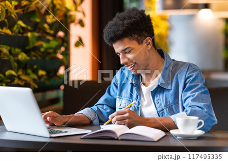 A smart African American young guy is seated at a table, working on a laptop while sipping from a cup of coffee. He appears focused and engaged in his work. 117495335