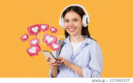 Smiling lady student in casual wear using her smartphone wearing white headphones, sending love message, carrying grey backpack, standing on yellow background 117495337