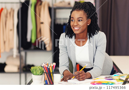 A smiling young African American woman sits at her desk, engrossed in designing new clothing pieces with colored pencils spread out before her 117495366