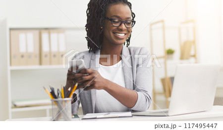 African American businesswoman smiling while using smartphone at her office desk, engaging in mobile communication. 117495447