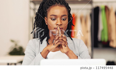 Pensive African American woman is sitting in front of a laptop computer, typing on the keyboard with a focused expression. The screen displays graphs and data, indicating work or research being done. 117495448