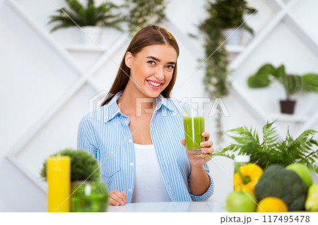 A woman smiles happily while holding a glass of green smoothie in her kitchen, surrounded by plants and fresh produce. A woman smiles happily while holding a glass of green smoothie in her kitchen, surrounded by plants and fresh produce. 117495478