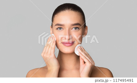 A woman is standing in front of a mirror, applying a facial cleanser to her face with her fingertips. She is gently massaging the cleanser into her skin, focusing on different areas A woman is standing in front of a mirror, applying a facial cleanser to her face with her fingertips. She is gently massaging the cleanser into her skin, focusing on different areas 117495491