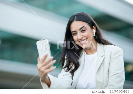 A young woman with dark hair, wearing a white t-shirt and a light blazer, stands in an urban setting. She smiles at the camera as she holds up her smartphone and appears to be using it. A young woman with dark hair, wearing a white t-shirt and a light blazer, stands in an urban setting. She smiles at the camera as she holds up her smartphone and appears to be using it. 117495493