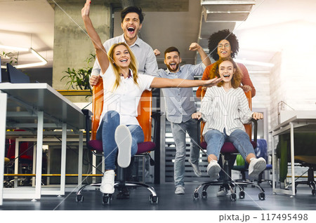A group of young office workers is caught in a moment of spontaneous joy, racing each other in office chairs. Their expressions are filled with laughter and excitement, suggesting a fun break A group of young office workers is caught in a moment of spontaneous joy, racing each other in office chairs. Their expressions are filled with laughter and excitement, suggesting a fun break 117495498